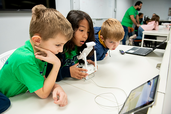 Image of young students working at a desk with iPad, laptop, and radio controller.