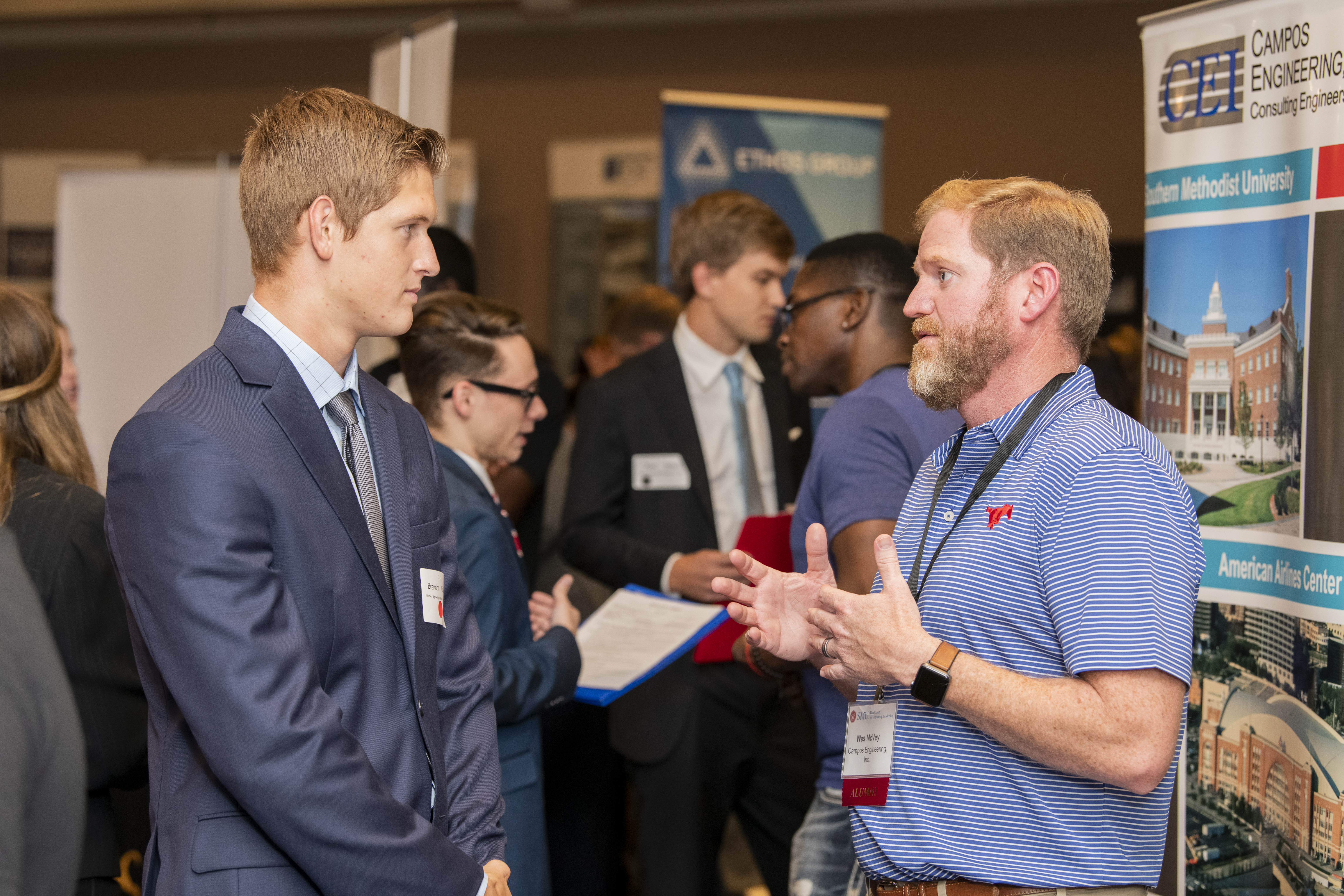 A mentor and a mentee engaged in conversation during a professional networking event, with other participants interacting in the background.