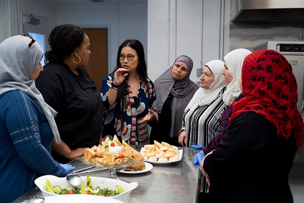 Founder Jin-Ya Huang speaking with a group of women in a kitchen setting with food on the counter representing break bread break borders.