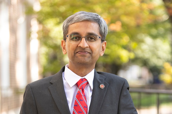 Dinesh Rajan in a dark suit and red tie stands outdoors, leaning against a railing with trees in the background.