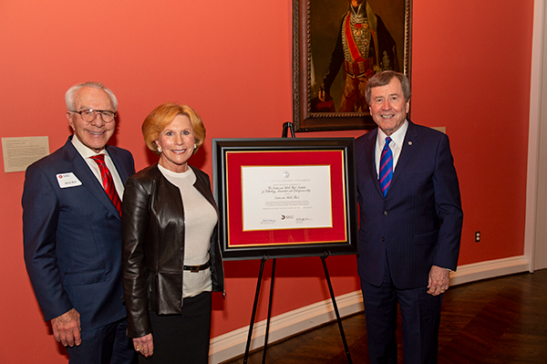 Three people posing with a framed certificate in front of a red wall.
