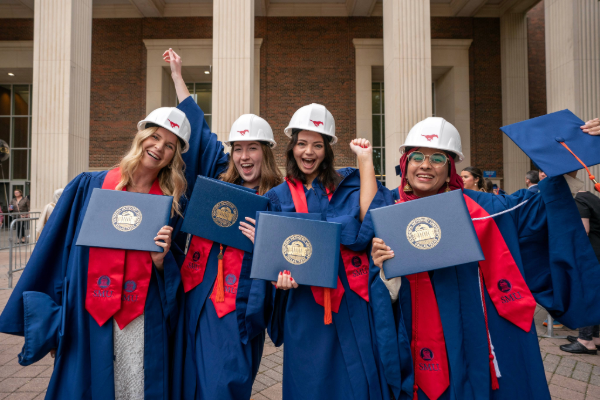 Students wearing SMU hard hats smile and hold up their diplomas on graduation day in their regalia
