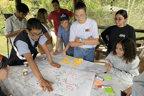Students stand around a table and observe a man draw a map of an area in Colombia