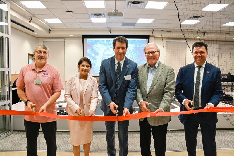 Faculty, executive board members and Quanser representatives stand with Dean Jalili and Dr. Bobby B. Lyle in front of the new lab. They are holding a red ribbon and about to cut it with scissors to celebrate the new lab's grand opening.