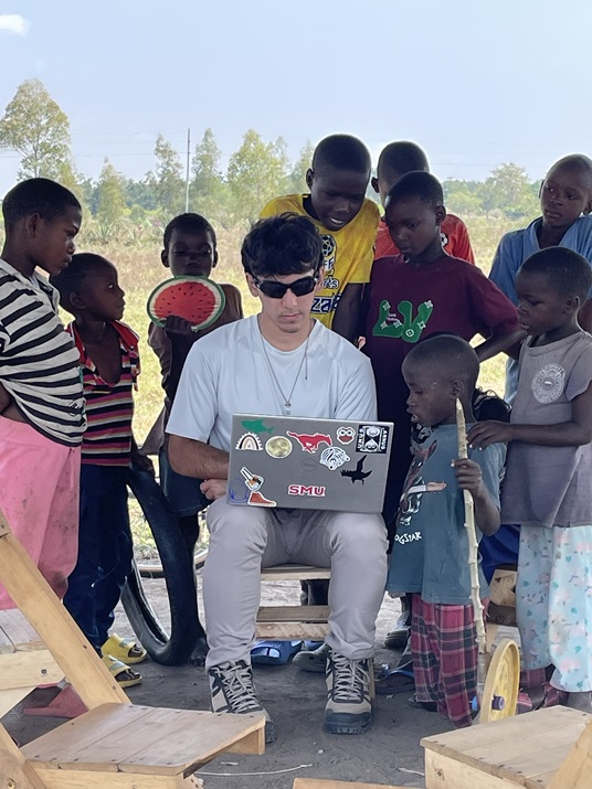 SMU Lyle student Kristian Perez sits on a chair with a computer on his lap, surrounded by small children who are watching him work in Tanzania.
