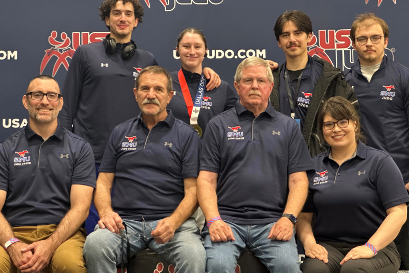 The 2025-26 SMU judo team and coaches pose for a group photo at a USA Judo event, with one student wearing a medal and smiling proudly. 