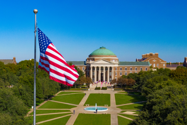 An ariel view of an American flag waving in front of a clear blue sky and Dallas Hall at SMU