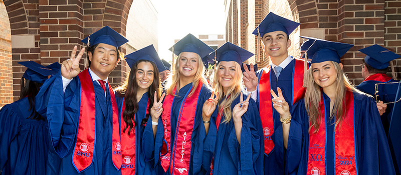 Group of six SMU graduates in blue caps and gowns with red stoles smiling and making the Mustang hand sign while standing outdoors near brick archways.