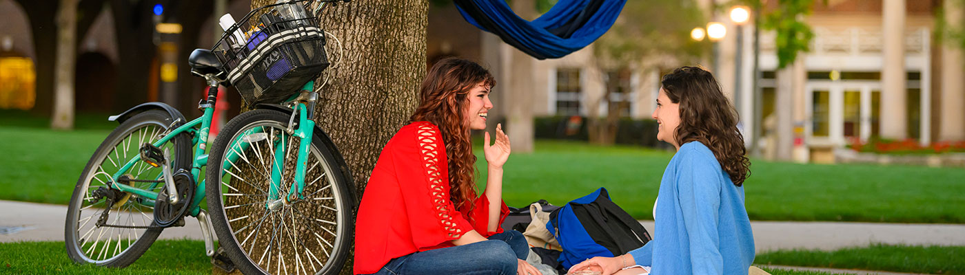 Two female students sit on the grass in front of a tree on a sunny day, with a bright blue bicycle parked beside them.