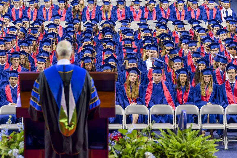 Dean Myers, from behind, in graduation robes speaking to 2025 graduates in robes