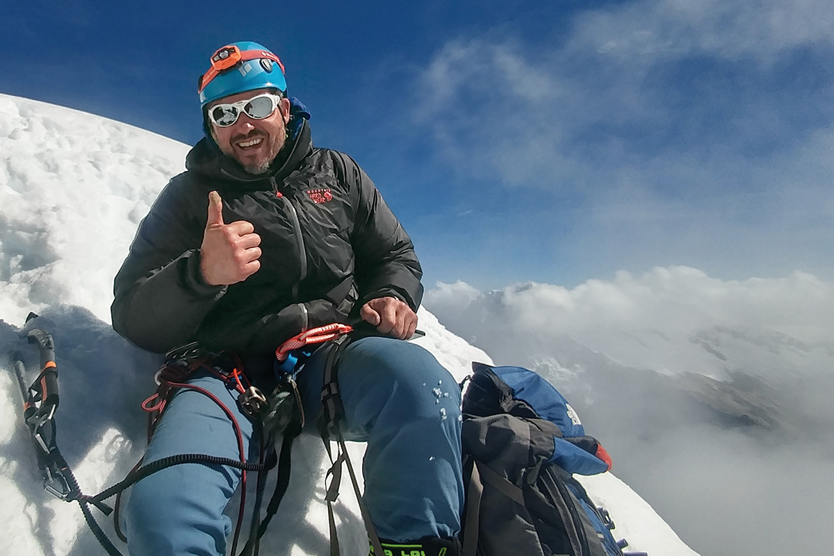 Dean Todd Milbourn gives a thumbs up sitting atop a snowy mountain peak