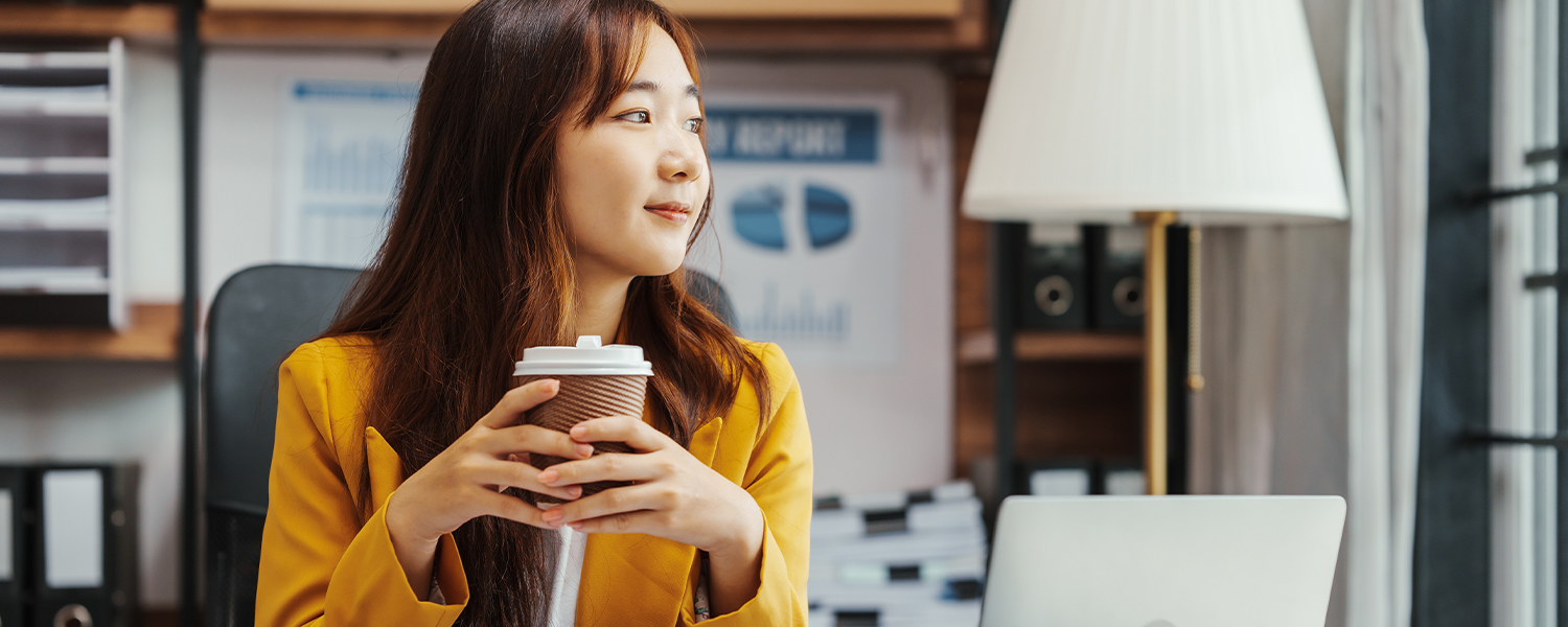 A woman drinking coffee and looking up from her laptop in thought.