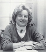 Betty Maynard wearing a white turtleneck and siting with her arms crossed on a table