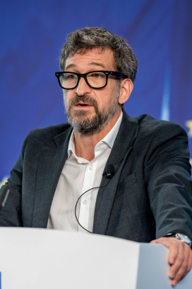 Middle-aged man with black blazer and white collared shirt with black framed glasses speaking at a podium. 