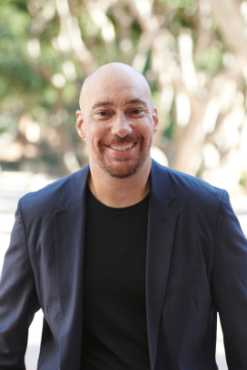 Adult male with glasses ,wearing a dark blazer and shirt, smiling against an outdoor background.
