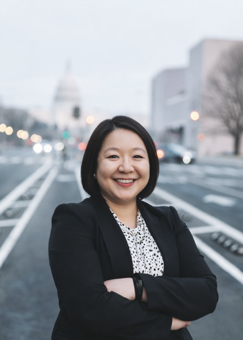 Adult Female in business attire. Black blazer with light colored bloused with a short bob hair cut and in a crossed arm stance against a city street background.