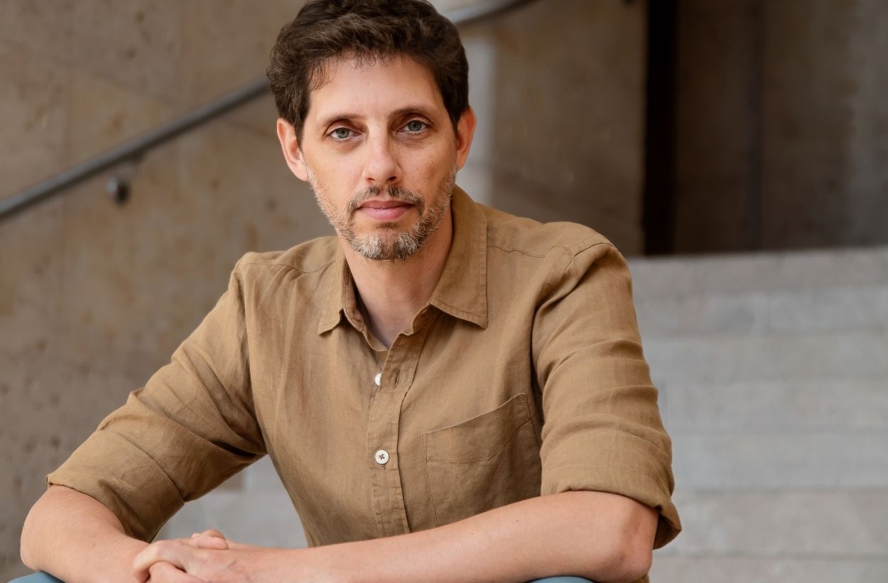 Professor Ariel Ron sitting on gray steps wearing a taupe shirt looking directly into the camera
