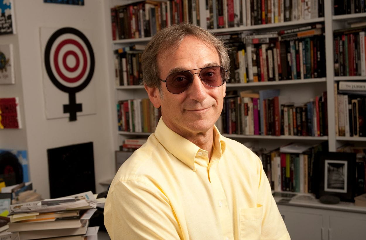 professor wearing yellow button up shirt with sleeves in an office in front of full bookshelves.