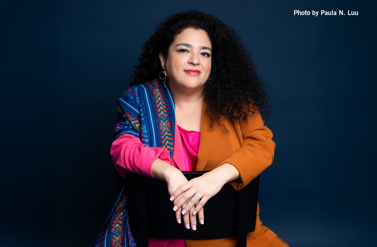 Marcela Fuentes sits backward on a chair, hands crossed at the chair's back, wearing a suit with orange on one side and pink and blues on the other.