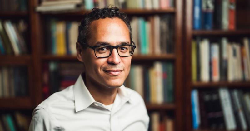 Picture of a man wearing black rimmed glasses indoors in front of a book case. He is wearing a white collared button up shirt.