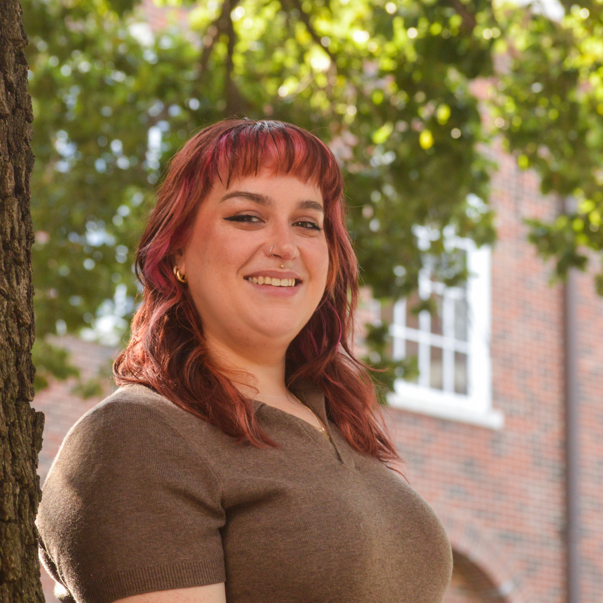 Sacoria Stixrud smiling outdoors beside a tree, with leafy greenery and a brick building softly blurred in the background.