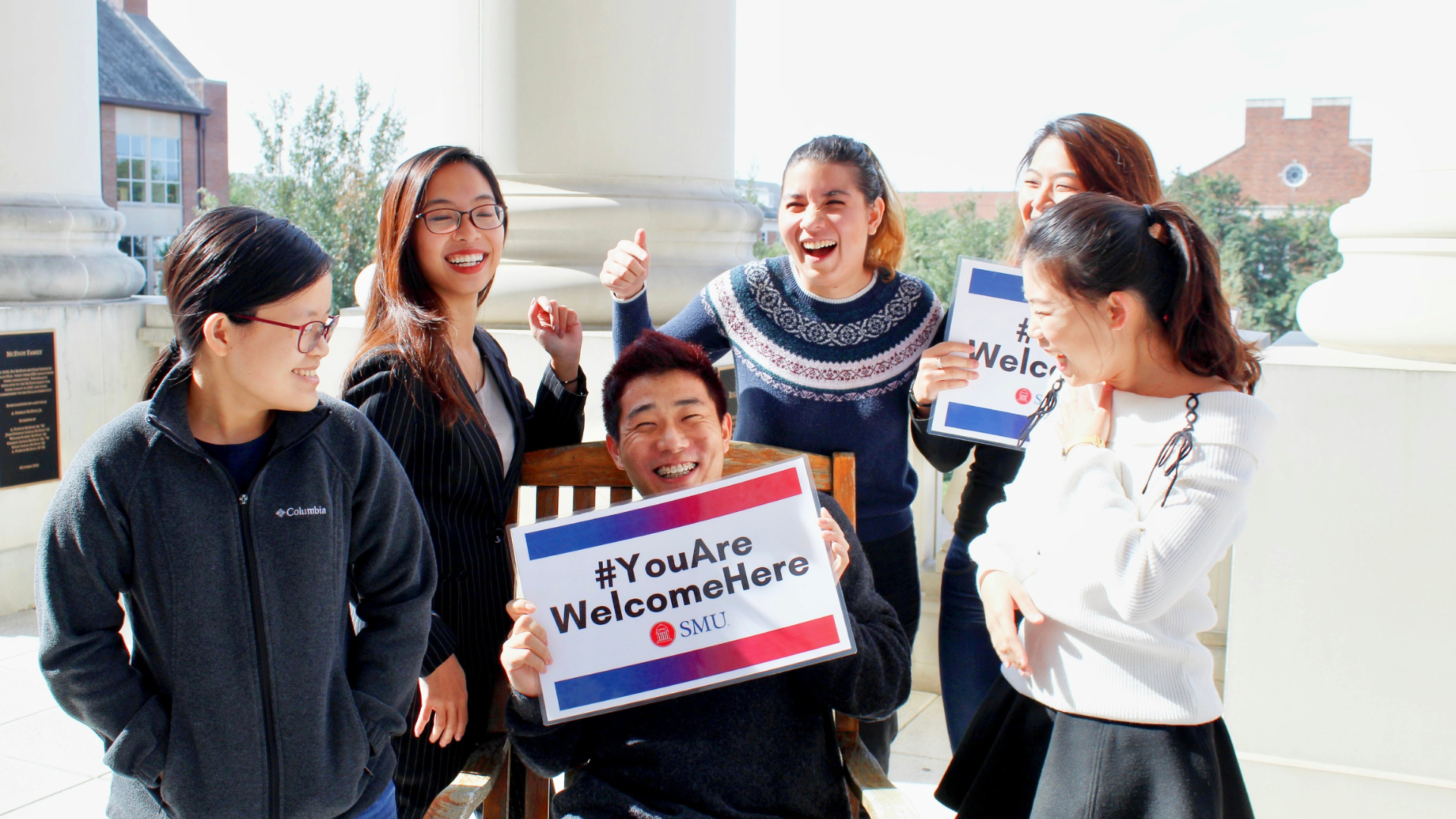 A male student sits in a wooden chair outside on a sunny day, holding a small sign that says You Are Welcome Here. Four female students are standing near the male student, and all are smiling and laughing.