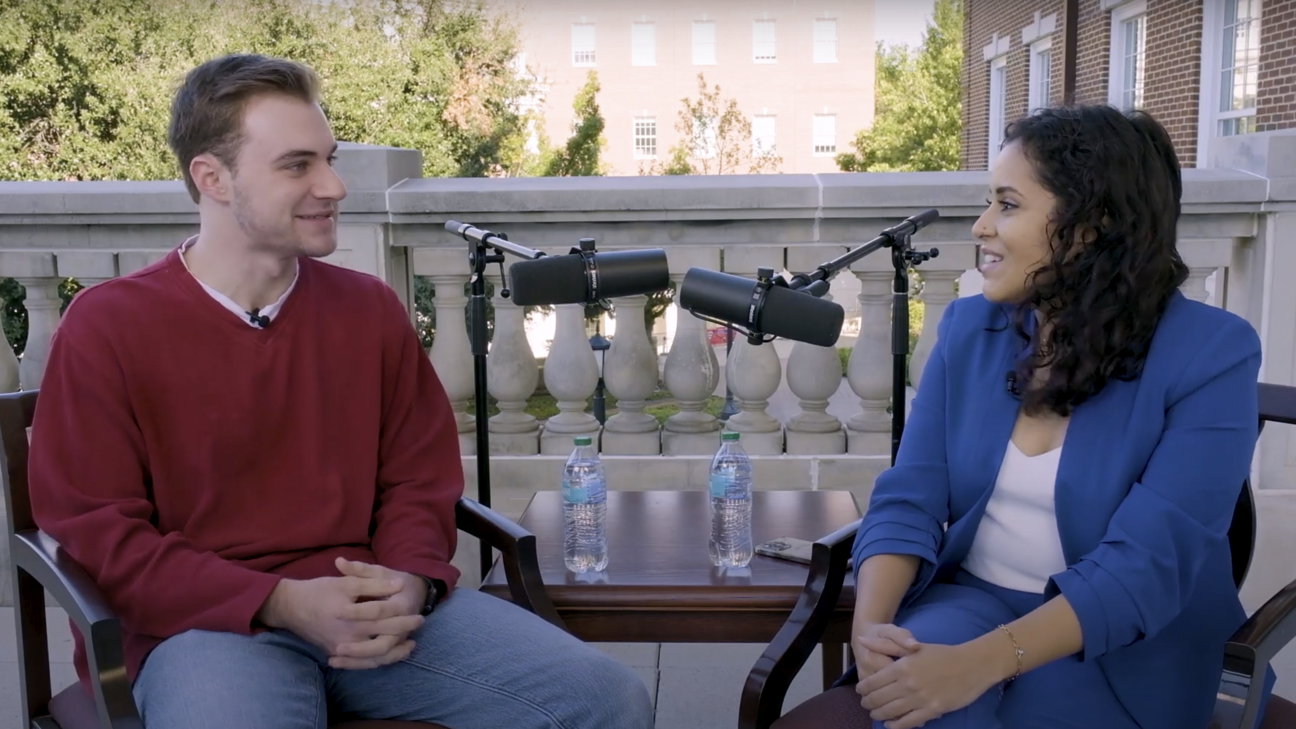 A male student in a red sweater and a female administrator in a blue blazer sit outdoors in front of microphones.