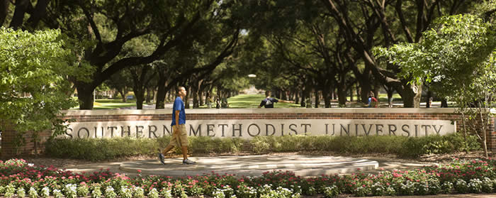 A man in tan cargo shorts and a blue shirt walks in front of the SMU sign on the tree-lined Boulevard. 