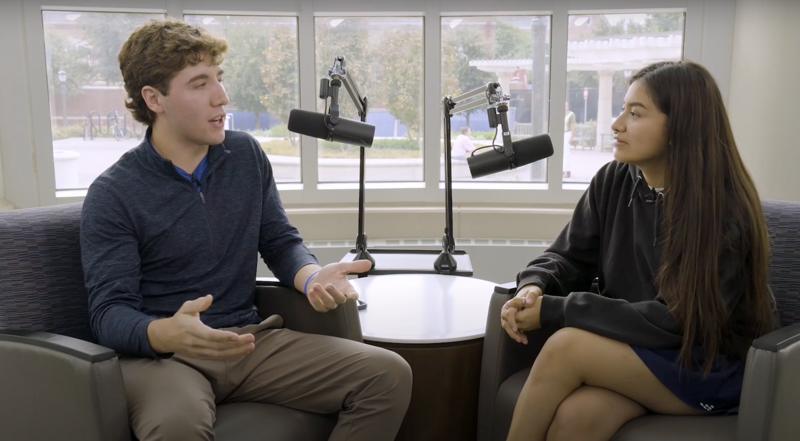 A male student speaking while gesturing with his hands, while a female student listens with an open expression. Both are sitting in chairs in front of microphones inside a well-lit room.