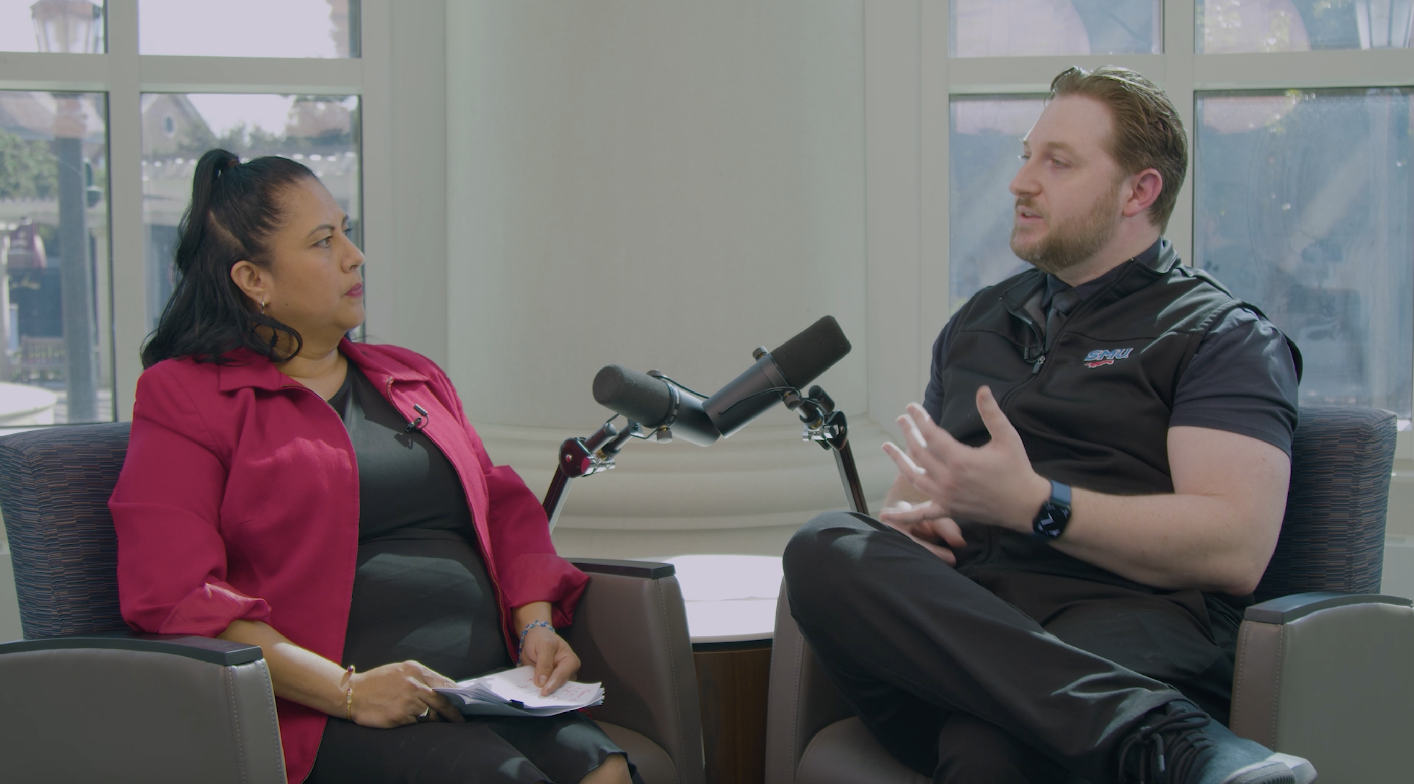 A woman in a dark pink blazer and a man in an SMU polo shirt sitting in chairs in front of microphones having a discussion