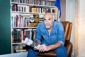 Robert Wilonsky sitting in a leather armchair holding a rolled-up newspaper with a bookshelf behind.