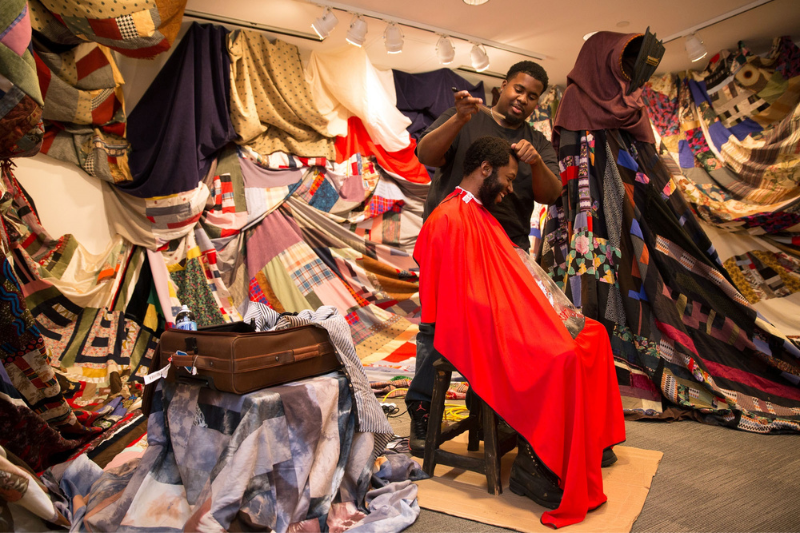 A man cuts another man's hair in front of many colorful fabrics hanging on the wall