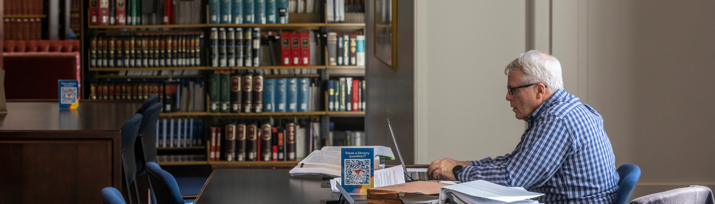 older man studies at a table with a laptop. books line the shelves behind him.