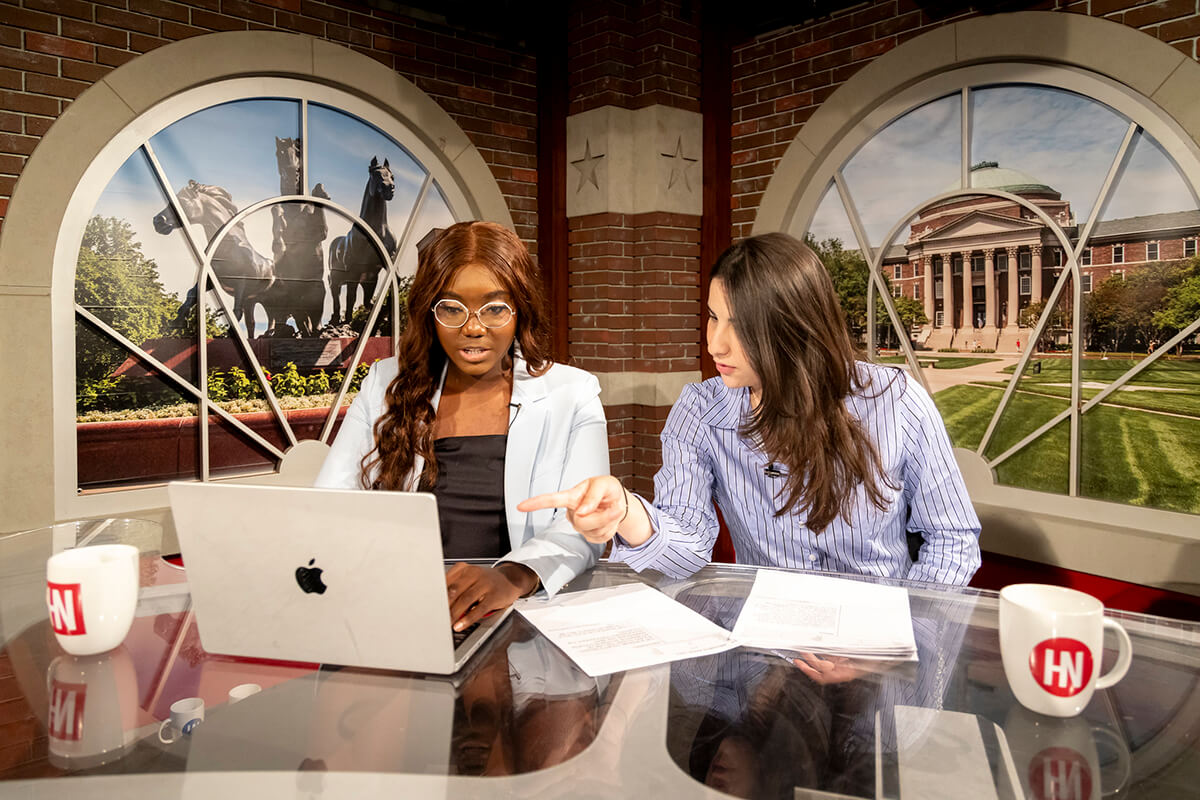 two female students sit at a mock news room desk