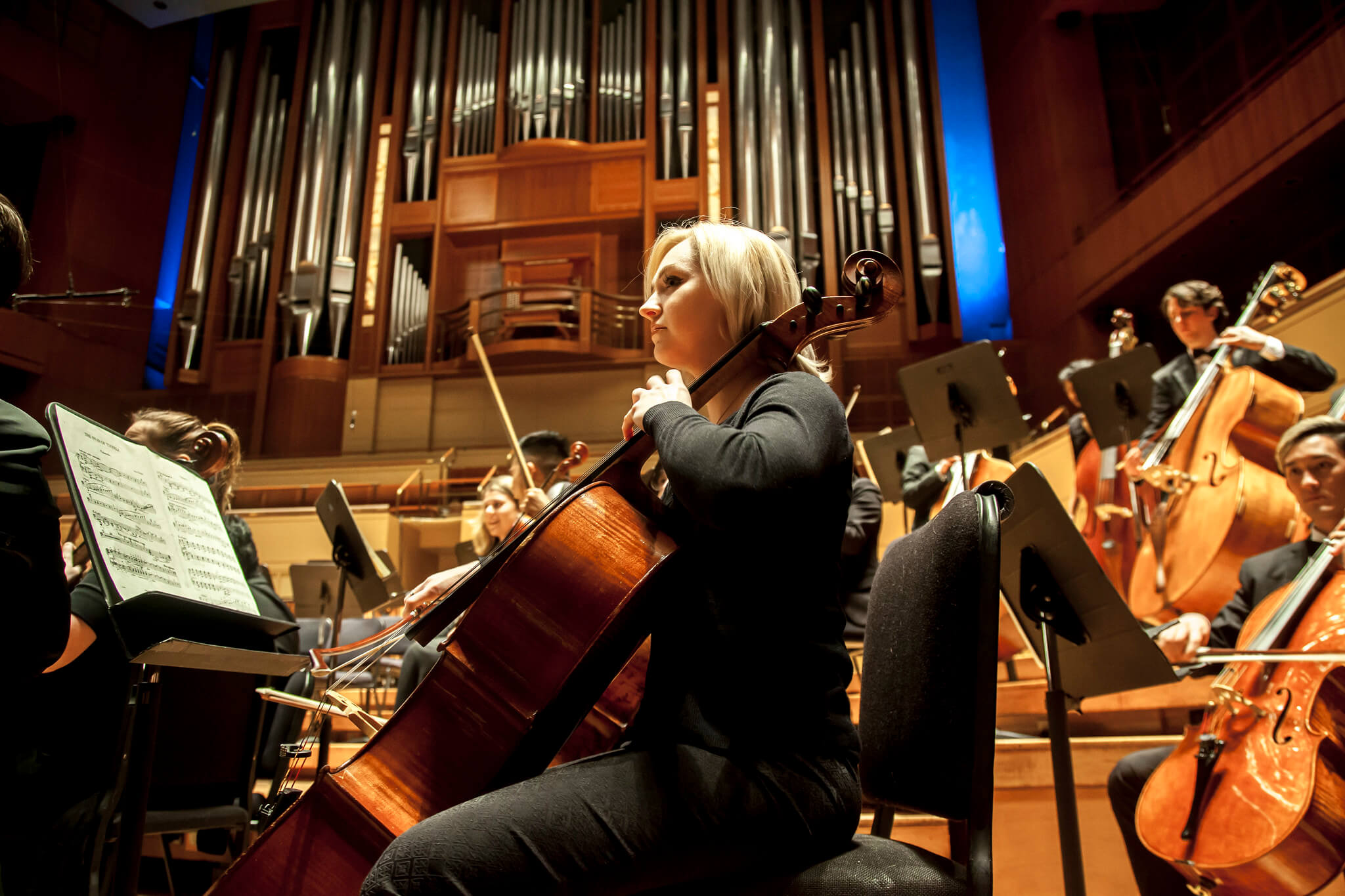 Orchestra on stage, girl playing cello with organ in background