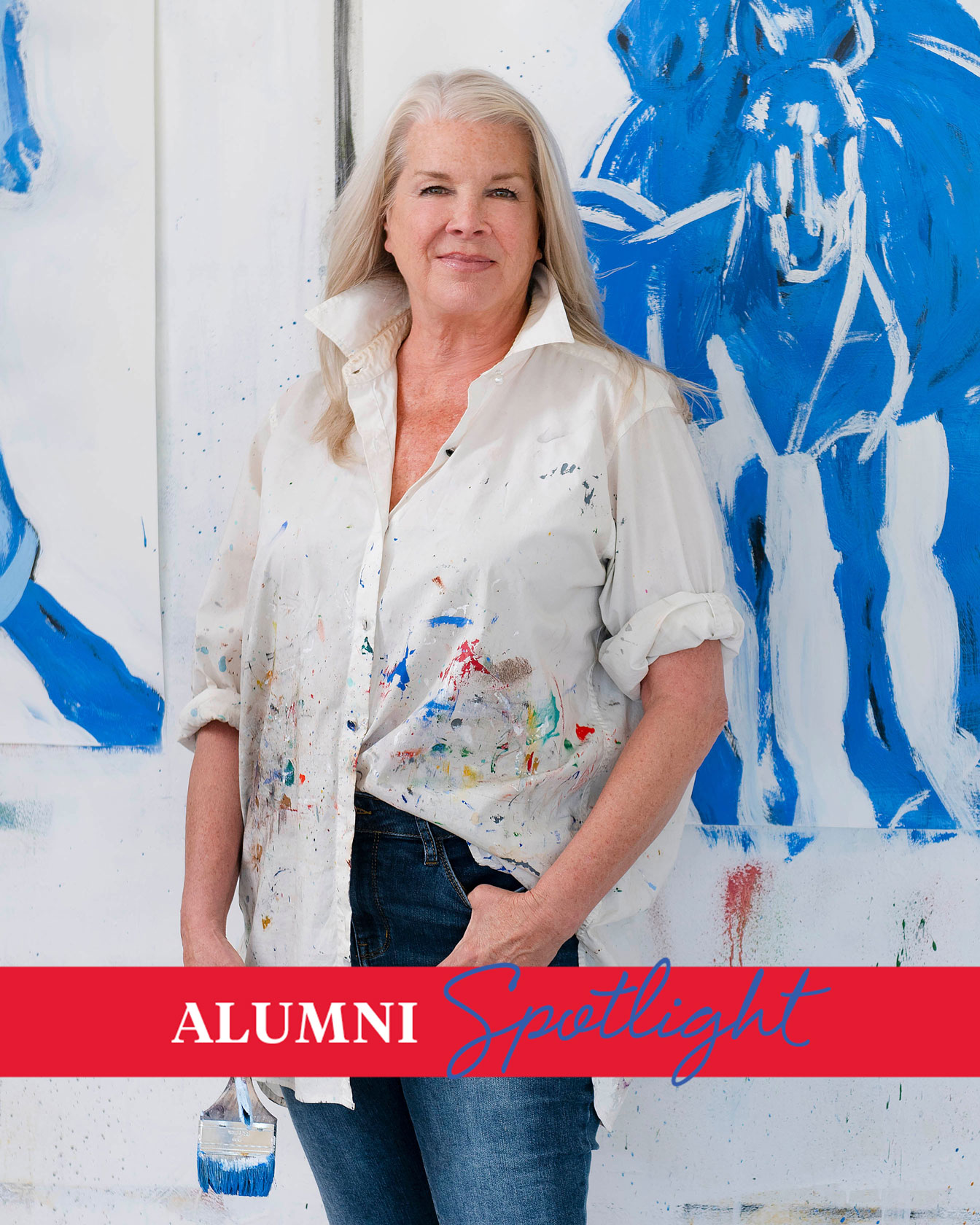 Division of Art alum Mel Auberty poses in front of canvases with blue horses holding a paintbrush