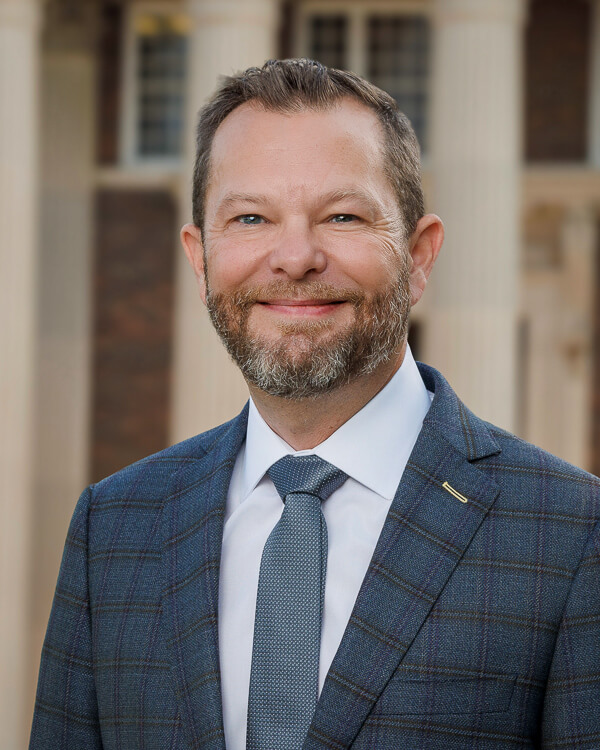 a man in a white shirt and plaid blue suit in front of a building with columns