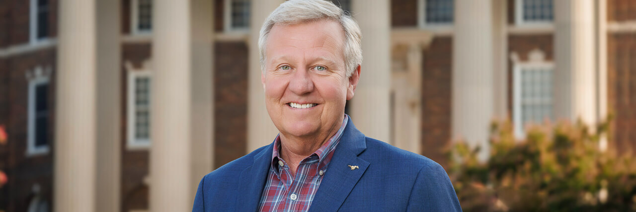 a man in a red plaid shirt and blue sports coat standing in front of a building with columns.