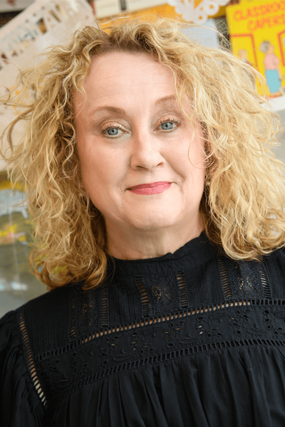 A woman with blonde curly hair wears a black blouse and smiles at the camera