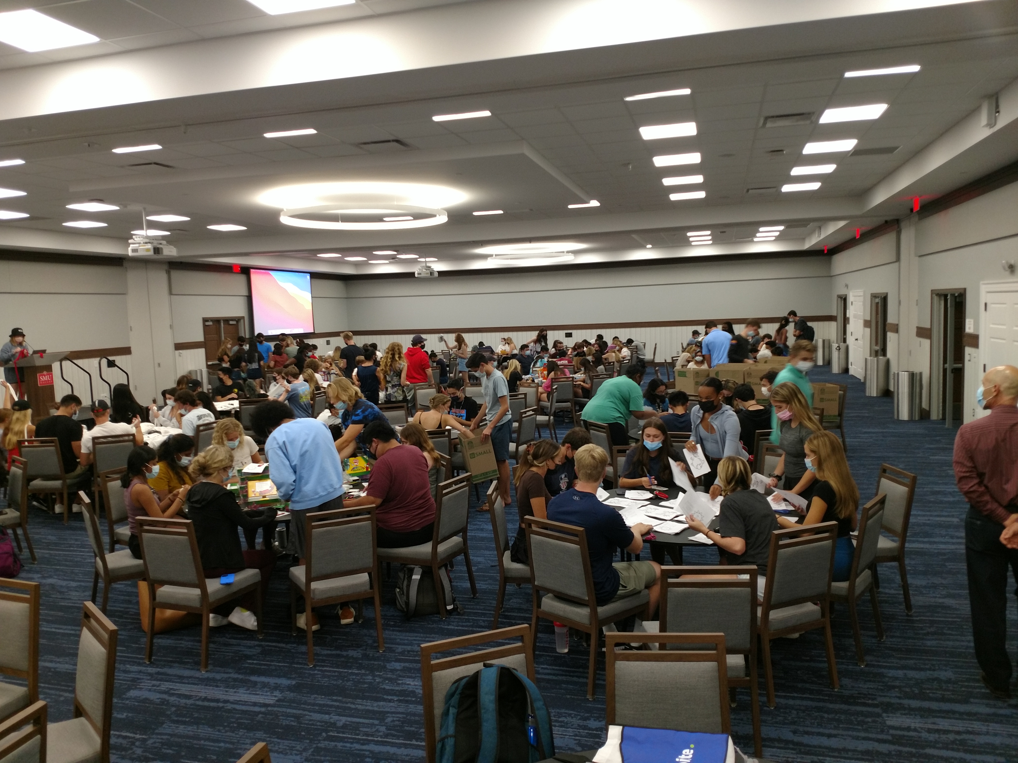 A group of 200+ students are seated around round tables. They are engaged in various activities for a school supply drive, such as putting together packets of activities and putting art supplies in an individual bag.