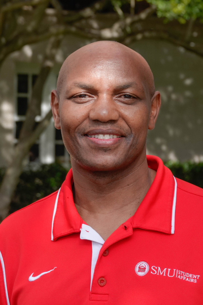 A professional headshot of SMU staff member Albert Mitugo standing outdoors, smiling confidently at the camera. He is wearing a red polo shirt with the logo of "SMU Student Affairs" and a white undershirt visible beneath the collar. The background features greenery and the side of a building, with trees providing some shade. 