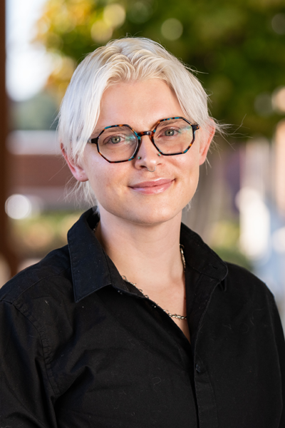 Headshot of Logan Swafford, a nonbinary staff member with light blonde, short hair parted down the middle and slight waves. Logan is wearing octagonal tortoiseshell glasses, small nostril and septum piercings, a silver necklace, and a long sleeve black button-up shirt. They have a gentle smile and a neutral, approachable expression. The blurred background features warm natural light in an outdoor setting.