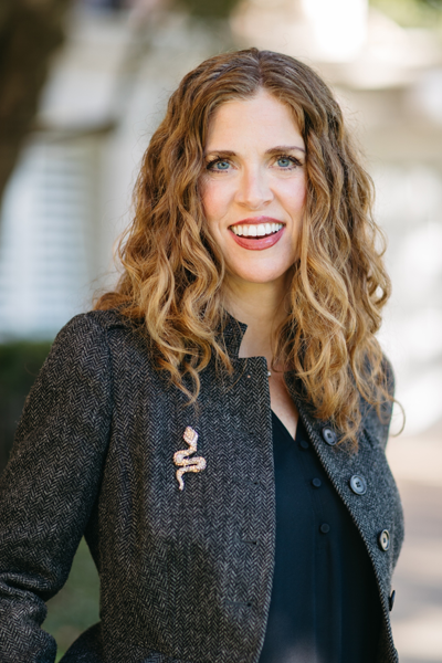  A professional woman with long, curly dark blonde hair smiles warmly at the camera. She is wearing a dark herringbone jacket adorned with a decorative snake brooch, over a black blouse. The background is softly blurred, suggesting an outdoor setting with natural light
