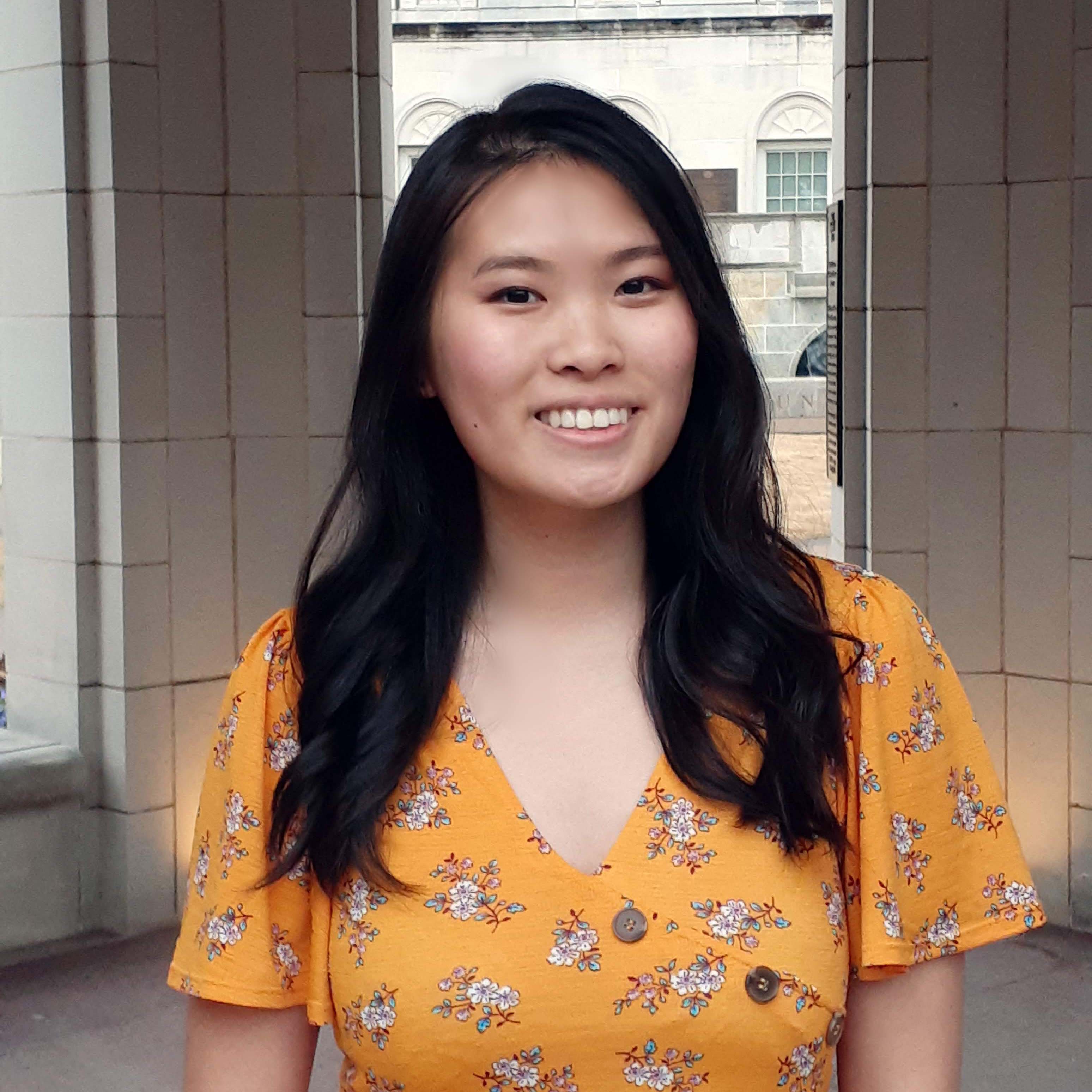 Tiffany Lam is an Asian American woman with long black hair past her shoulders. She wears a yellow dress with white flowers