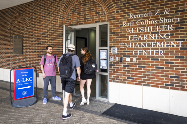A brick building with the words Kenneth Z. & Ruth Collins Sharp; Altshuler Learning Enhancement Center. An individual is holding the door open for two other people entering the building. To the left is a sign: "Welcome to the A-LEC" with an arrow pointing towards the entrance. 