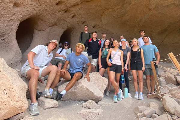 A group of young adults pose and smile inside a rocky cave-like opening, some sitting on large boulders and others standing behind them, dressed in casual outdoor clothing with water bottles and day-hiking gear visible among the rocks.
