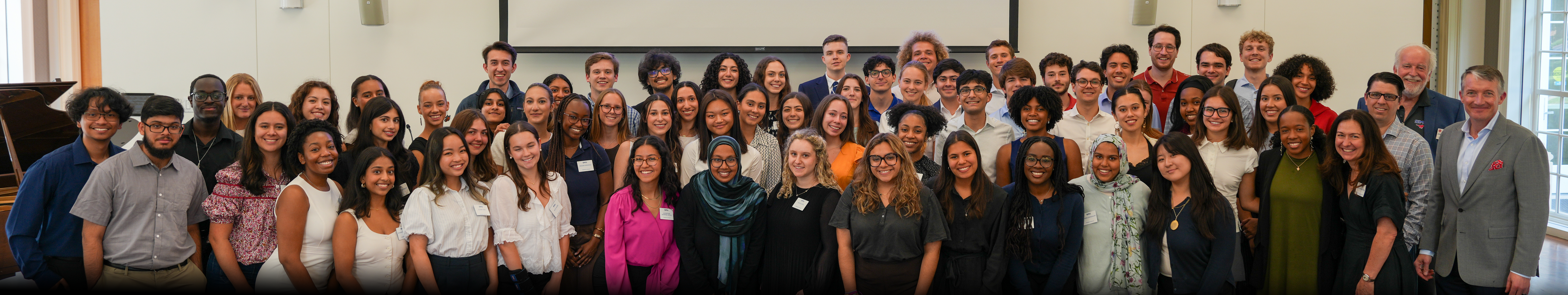 A collective photo of President's Scholars and staff pose together in Prothro Great Hall event space, standing in several rows in front of a projection screen.