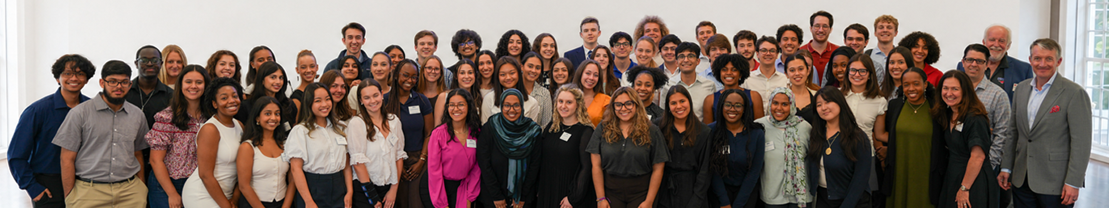 A collective photo of President's Scholars and staff pose together in Prothro Great Hall event space, standing in several rows in front of white wall
