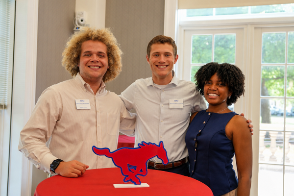 Three people stand close together behind a tall cocktail table with a red table cloth at an indoor event, smiling toward the camera; a red with blue mustang table decoration sit on the table in front of them, with large windows and neutral-colored walls in the background.