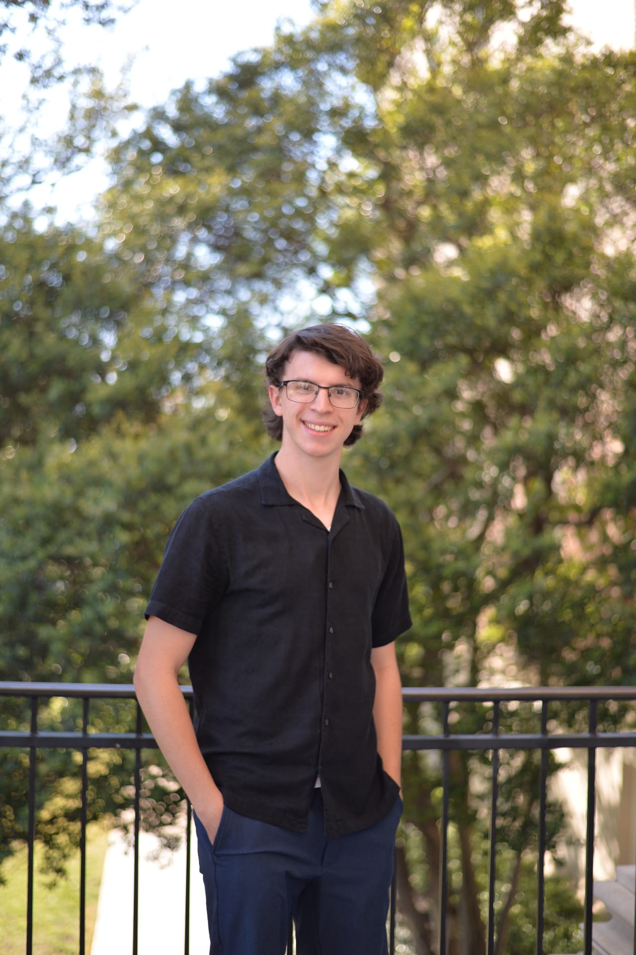 Joshua Ange is a white man with brown hair. He wears glasses, a black short sleeve dress shirt, and navy blue slacks. He stands in front of a black iron fence with trees in the background.
